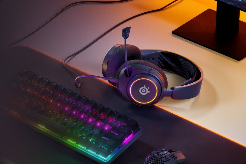 An image of Arctis Nova headset laying on the desk, accompanied with glowing keyboard and mouse.